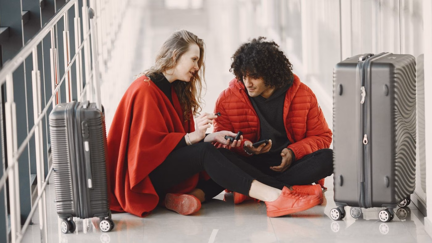 A young couple sits on the floor at an airport, engaging and relaxed, with suitcases around them.