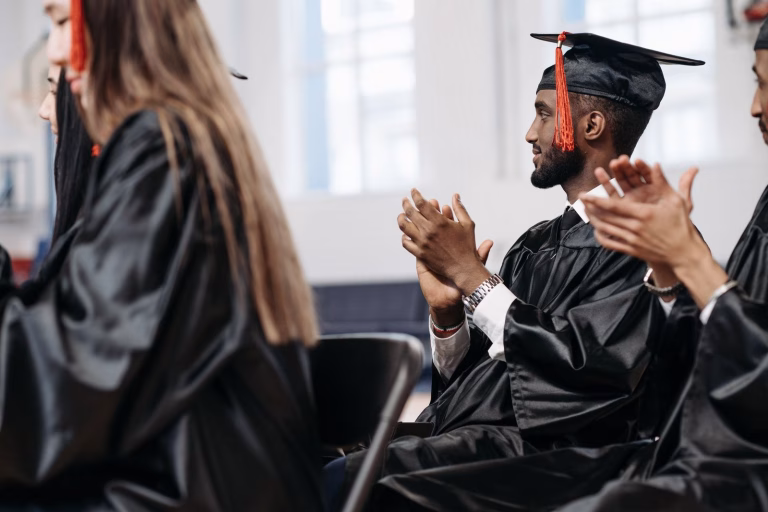 Graduates in caps and gowns clapping during a joyful graduation ceremony indoors.